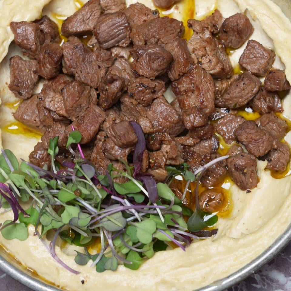 Hummus with seasoned beef chunks and microgreens in a metal pan on a marble surface.