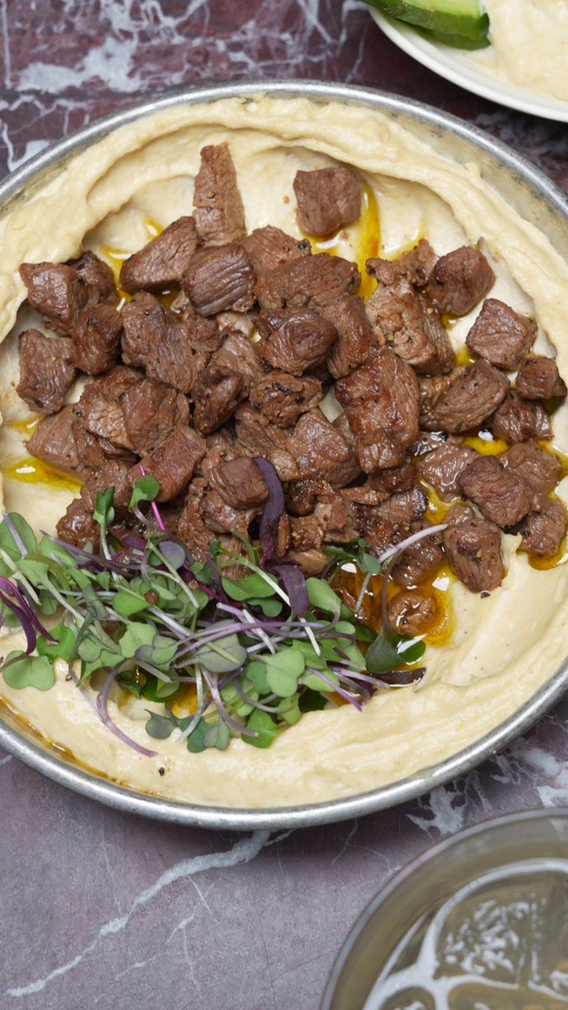 Hummus with seasoned beef chunks and microgreens in a metal pan on a marble surface.