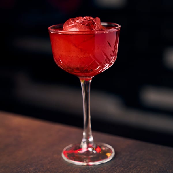 Red cocktail in a stemmed glass with a red ice sphere on a dark bar.