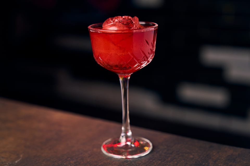 Red cocktail in a cut glass coupe with a large ice sphere, on a dark bar.