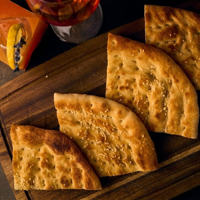 Four pieces of sesame flatbread on a wooden board with cocktails in the background.