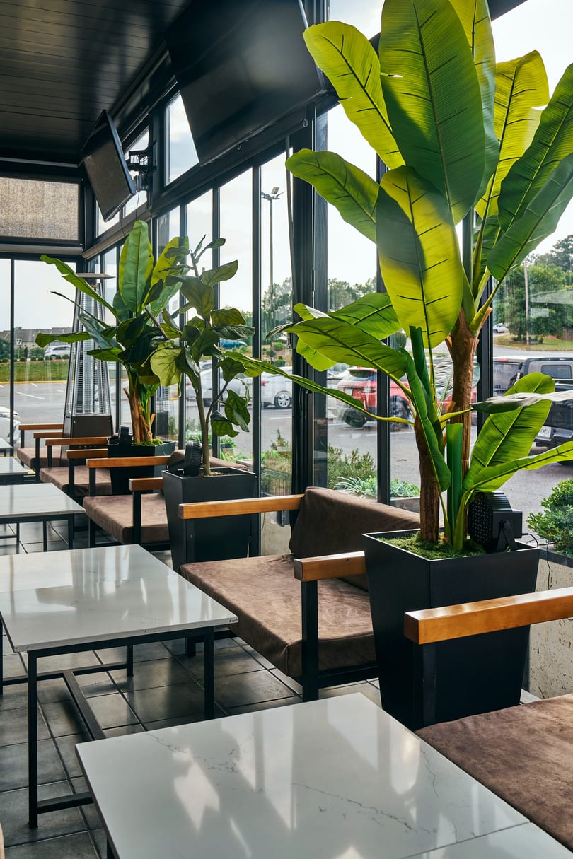 Modern restaurant interior with white tables, brown benches, large green plants, and expansive windows.