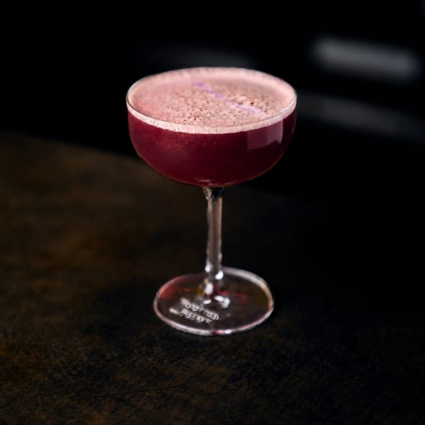 Foamy deep red cocktail in a Woodford Reserve coupe glass on a dark table.
