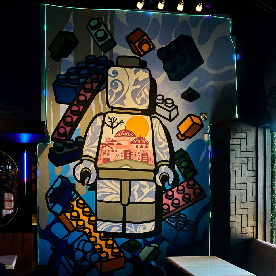 Colorful mural of a stylized robot surrounded by geometric shapes decorates a dimly lit restaurant dining area with tables and chairs.