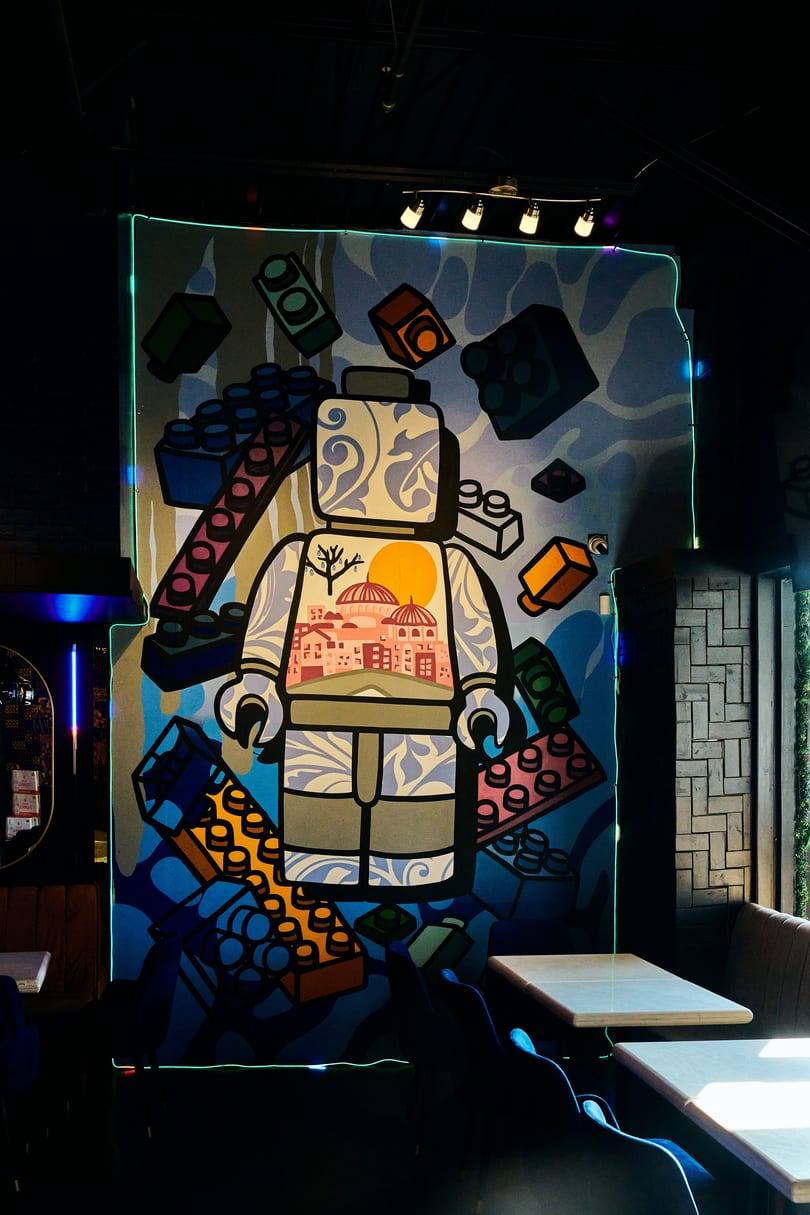 Large colorful mural of a Lego-like figure with a cityscape and sun, surrounded by bricks, in a restaurant.
