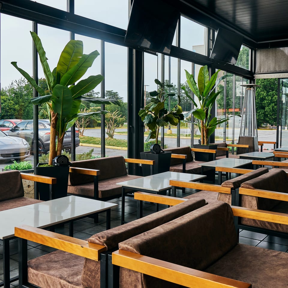 Modern restaurant interior with brown lounge seating, white tables, tropical plants, and floor-to-ceiling windows.