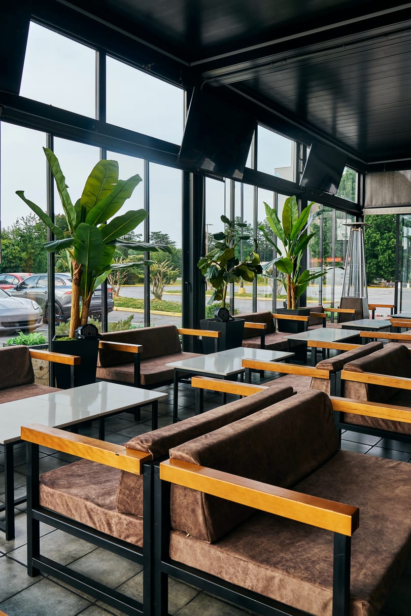 Spacious restaurant patio with brown cushioned sofas, white tables, large windows, and potted plants.