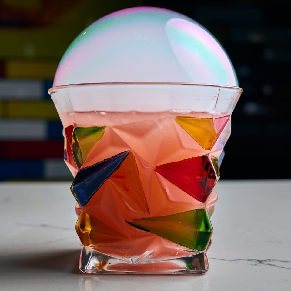 Pink cocktail in a colorful geometric glass, topped with a shimmering smoke bubble.