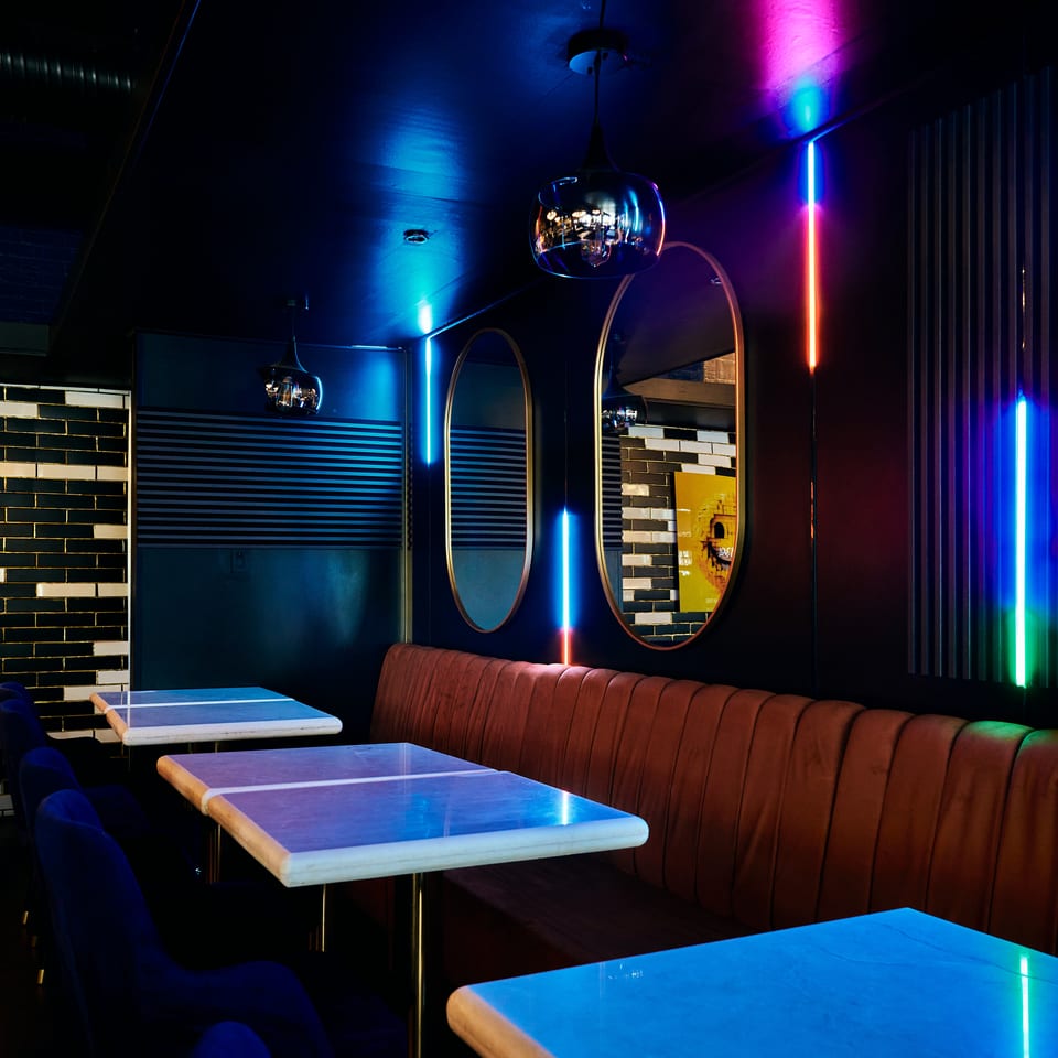Moody restaurant interior with blue-lit tables, velvet seating, pendant lamps, mirrors, and colorful neon accents.