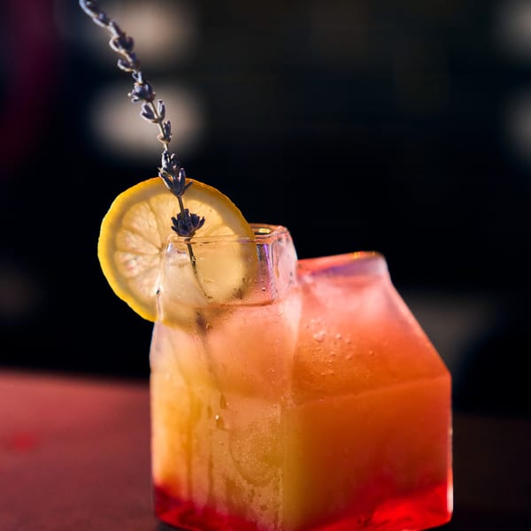 Vibrant orange and red cocktail in a unique glass, garnished with lemon and lavender.