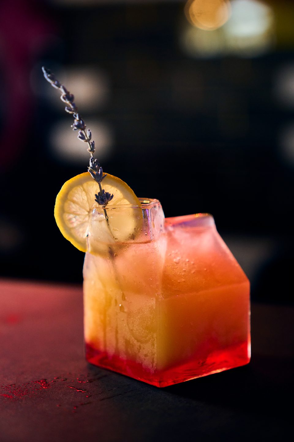 A vibrant orange and red cocktail in a unique glass, garnished with a lemon slice and lavender sprig.