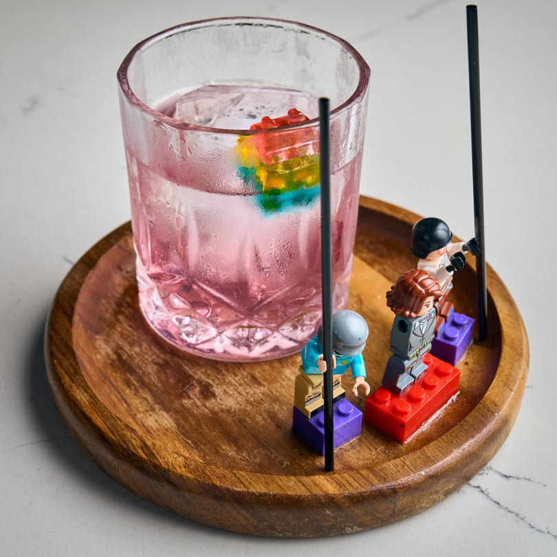 Pink cocktail with colorful ice cubes and Lego figures on a wooden tray.