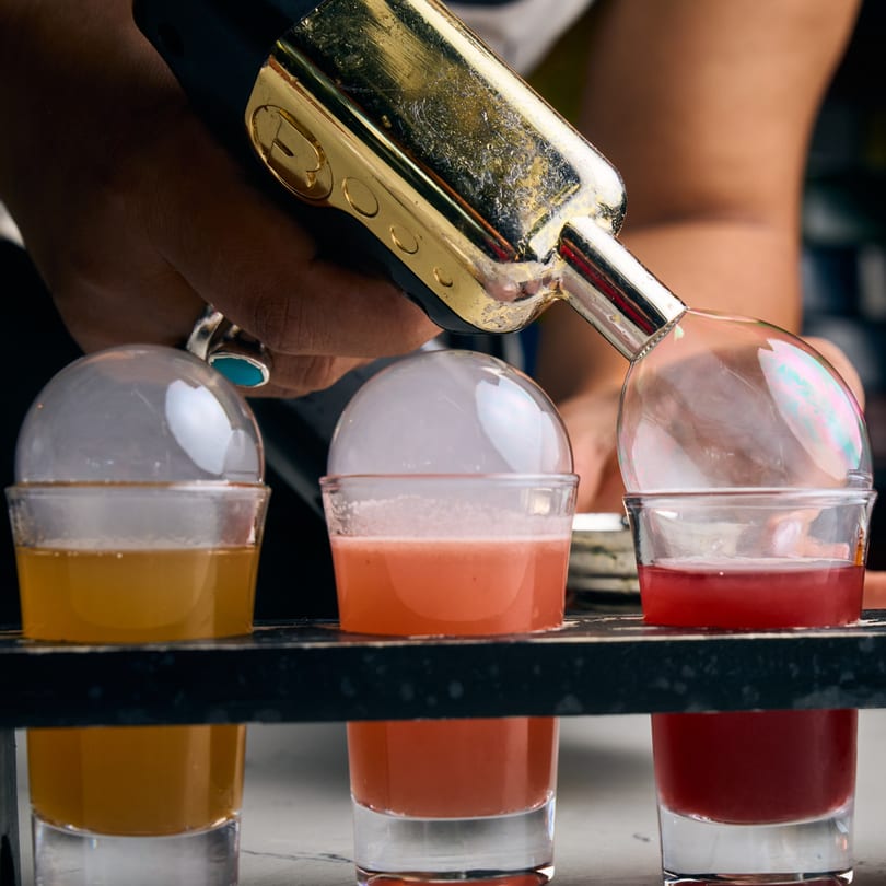 Bartender using a torch to create smoke bubbles on three colorful Adana Restaurant cocktails.